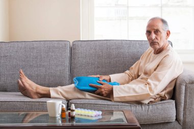 Son sınıf öğrencisi kanepeye oturmuş sıcak su torbasıyla dizlerine masaj yapıyor. (Sağlık ve zindelik)