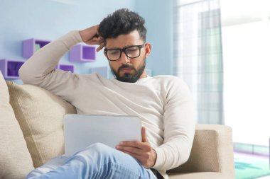 Genç adam evdeki kanepede dinleniyor ve tabletini kullanıyor..