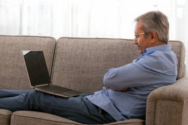 Evde oturup dizüstü bilgisayara bakan yaşlı bir adamın portresi..