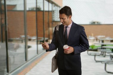 İş adamı resmi giysiler içinde elinde kahve fincanı taşıyan ve cep telefonu kullanan biri.