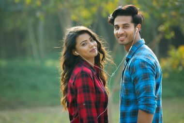 Bahçede dikilen genç çift bir kulaklığı paylaşıyor ve müzik dinliyor..