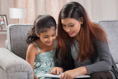 Anne evdeki kanepede oturan kızına kitap okuyor.