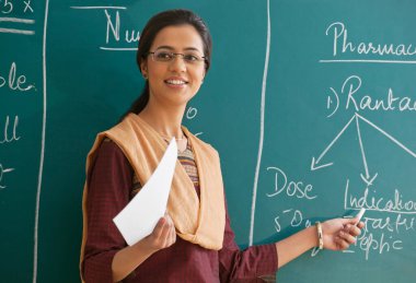 Kadın öğretmen tahtaya karşı durarak bir dersi açıklıyor