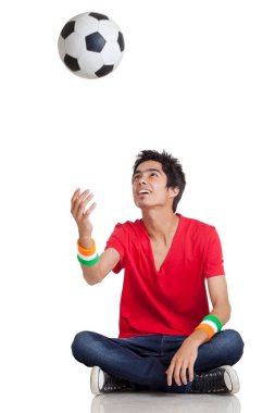 Genç bir çocuk, beyaz arka planda çapraz otururken futbol topunu atarken gündelik kıyafetlerini giyiyor.