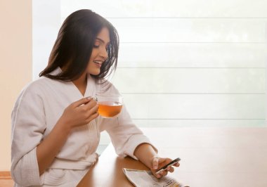 Yemek masasında çay fincanı tutarken mutlu genç bir kadın mesaj atıyor.