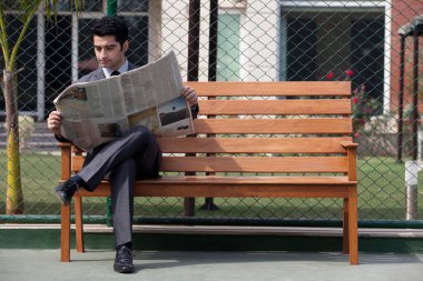 Genç erkek işletme yöneticisi tenis kortunda bankta otururken gazete okuyor.