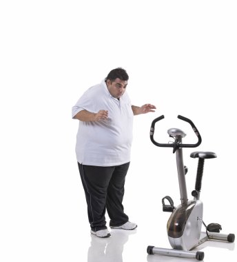 Şok olmuş obez bir adamın beyaz arka planda egzersiz bisikletine bakması.