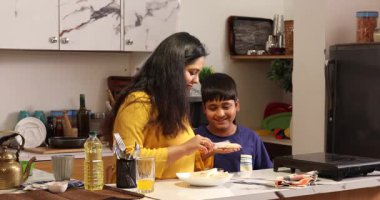 Anne sandviç yapıyor ve oğul mutfağı temizliyor.