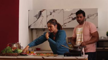 Kadın mutfakta dikilirken, adam tepsiden tabağa yemek servisi yapıyor..