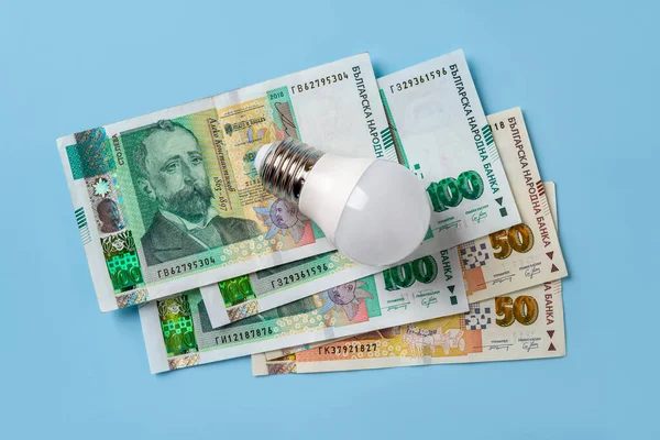 Light bulb on bulgarian lev banknotes over blue background. High energy costs concept. Power price rising in Europe. Soaring of electricity bills. Energy price spike. Top view.