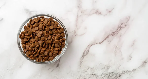 Dry pet food in a glass bowl over marble background. Glass feeding bowl full of gluten free dry protein kibbles for cats closeup. Complete food for domestic animals concept. Copy space. Top view.