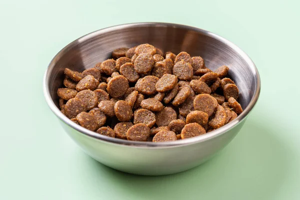 Dry pet food in a metal bowl over green background. Gluten free dry protein kibbles for cats in a stainless steel feeding bowl closeup. Complete dry food for domestic animals concept. Front view.