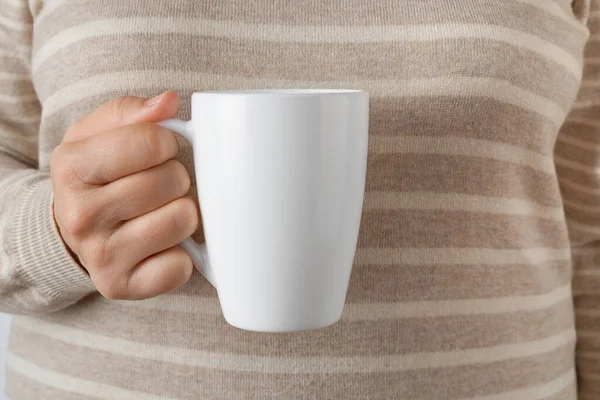 Woman hand holding white coffee mug. Female hand holds blank tea cup against striped wool sweater. Modern tableware, white crockery, hot beverage concept. Front view.