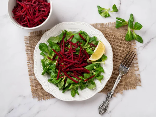 stock image Vegan salad of lambs lettuce and raw beet. Fresh cornsalad leaves, grated raw beet and lemon slice on a white plate over marble surface. Vitamin vegetable salad for low calories slimming diet. Top view.