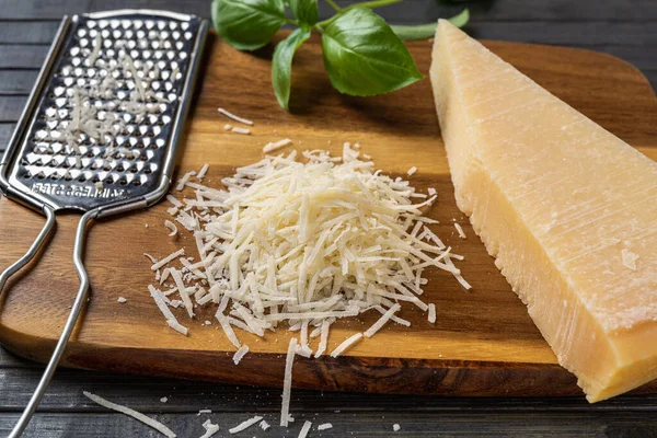Heap of grated parmesan cheese closeup. Tasty grana padano cheese whole and grated and stainless steel grater on a wooden cutting board. Italian dairy product. Hard cheese. Front view.