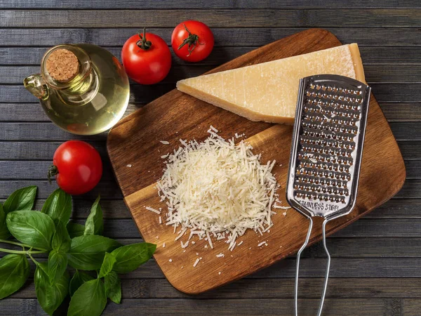 Parmesan cheese and grater on a cutting board. Whole and grated grana padano cheese, grater, olive oil, green basil and tomatoes over wooden background. Delicious dairy product. Top view.
