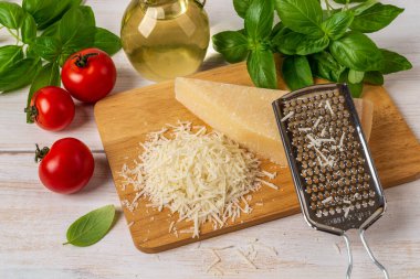 Kesme tahtasında parmesan ve el rendesi. Lezzetli sert peynir grana padano, paslanmaz çelik rendesi, kırmızı domates, zeytinyağı ve yeşil fesleğen otu beyaz ahşap masa üzerinde. Süt ürünü. Üst görünüm.