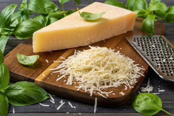 Shredded parmesan cheese and grater on a cutting board. Grana padano cheese whole wedge and grated, stainless steel grater and green basil herb over wooden background. Hard cheese. Front view.