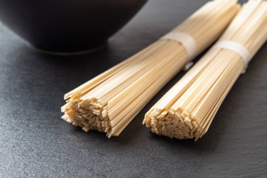 Raw udon nudles are ready for cooking. Two bunches of dried japanese vegan pasta from wheat flour and black bowl over black slate stone surface. Asian cuisine concept. Front view.