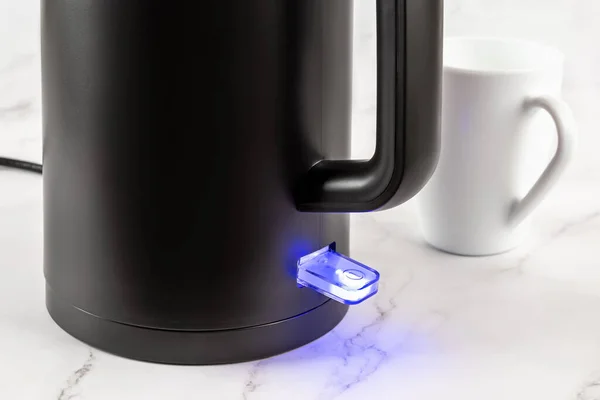 Electric kettle switch glows blue while turned on. Matte black kettle for heating water to boiling for tea and coffee. Modern class 1 electrical appliances for kitchen concept. Closeup.