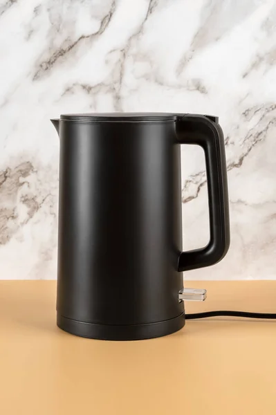 Matte black kettle against marble wall. Stylish electric kettle for heating water to boiling for tea and coffee. Class 1 electrical appliances for kitchen. Vertical frame. Front view.