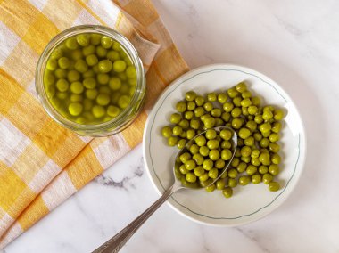 Mermer tezgahın üzerinde marine edilmiş organik bezelyeler. Cam kavanozun yanında konserve yeşil bezelye ve kaşık. Sebze proteini, salata malzemesi konsepti. Üst görünüm.
