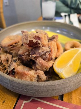 Çıtır kalamar, parlak bir limon kaması eşliğinde yuvarlak bir kasede ustalıkla düzenlenir ve altın rengini sıcak ahşap bir masada öne çıkarır. Misafirler canlı bir yemek atmosferinin keyfini çıkarıyorlar..