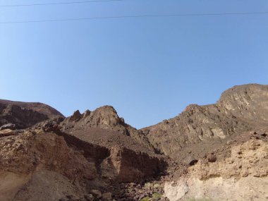 Kahverengi kayalık arazili engebeli dağların nefes kesici manzarası. Manzara, uzak bir çölde sakin bir atmosfer yaratan parlak mavi gökyüzünün altında doğal güzellikleri sergiliyor..