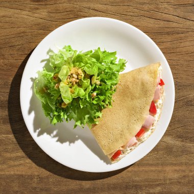 Mayonezli krep, jambon ve domates, yanında salata. Tahta bir masada, beyaz bir tabakta.