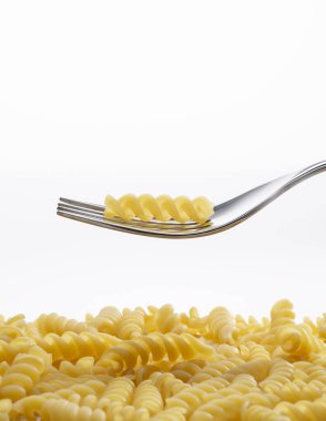 Çelik çatal üzerinde çiğ fusilli makarna avuç içi ve sarı fusilli yüzey üzerinde yüzüyor, temiz arka planda izole edilmiş.