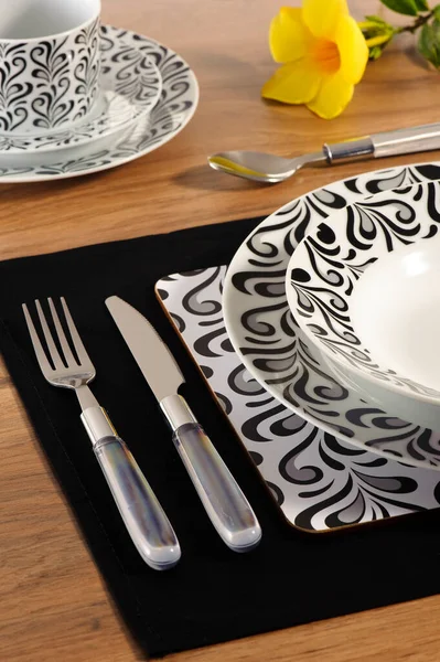 Table set up with dinnerware and silverware, ready for a meal. Close up of the fork and knife, with a set of dishes aside on a black table mat, and cup and saucer on the background.