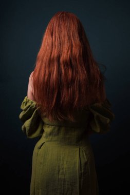 Omuzundan kayan yeşil bir elbise giyen, uzun kızıl saçlı gizemli bir kadının arka görüntüsü.