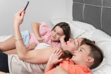 Genç anne ve tatlı kızı ve oğlu akıllı telefonlarıyla selfie çekiyorlar ve evde yatakta yatarken gülümsüyorlar. Yüksek kalite fotoğraf