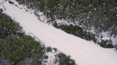 Karlı bir dağ beldesindeki bir teleferiğin hava aracı görüntüsü. Sahne, dağlık bir çevrede kışın manzaraları, çam ağaçları ve karla kaplı yamaçların üzerindeki kablo boyunca sandalyeleri gösteriyor. Yüksek kalite 4k görüntü