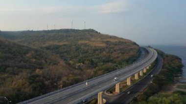 Uzakta bir sahil şeridi yakınında rüzgar türbinleri olan tepelik, ormanlık bir arazide esen bir otoyolun havadan görüntüsü..