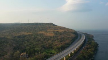 Uzakta bir sahil şeridi yakınında rüzgar türbinleri olan tepelik, ormanlık bir arazide esen bir otoyolun havadan görüntüsü..