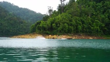 Yüksek yemyeşil dağlar ve yoğun tropikal yağmur ormanlarıyla çevrili, dingin bir göl boyunca süzülen geleneksel uzun kuyruklu bir teknenin manzaralı geniş bir görüntüsü. Kayık sakin sularda hafif dalgalanmalar yaratır kayalık bir kıyıdan geçerken. 