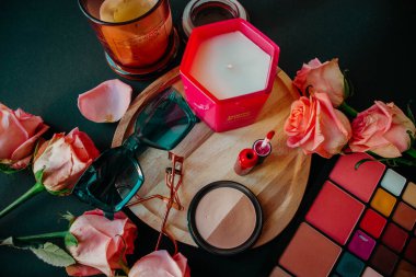 Candle on a wooden stand with flowers, glasses, lipstick and a palette with eye shadows on a black background