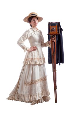Kameralı genç bir kadın bej renkli 1880 'lerden kalma bir elbise ve saman şapka giyiyor.