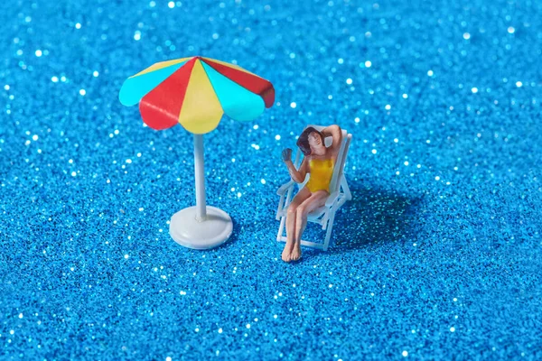 stock image High angle of miniature plastic lady sitting on chair under bright colorful umbrella on blue sparkling surface