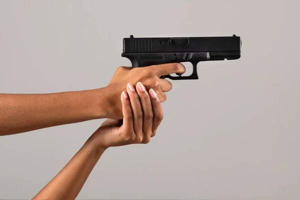 Crop anonymous African American woman with black gun pulling trigger on gray background