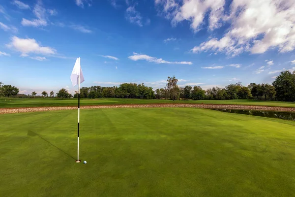 Picturesque view of golf course with flag pin and ball on green lawn under blue sky on sunny day