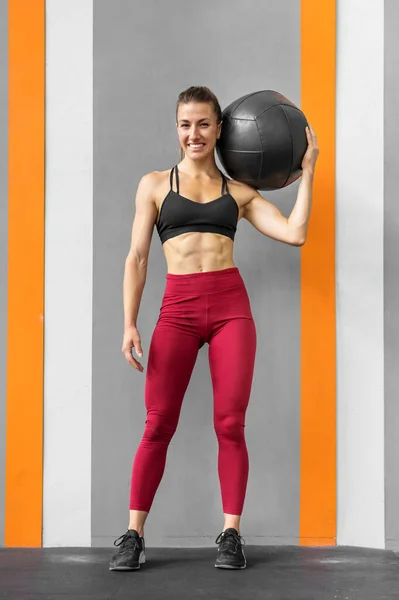 Full body of positive young muscular female athlete with long dark hair in sportswear smiling and looking at camera while standing in gym with medicine ball in hand
