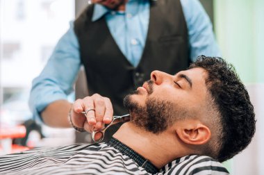 Crop anonymous barber using scissors while cutting beard of male client in professional barbershop