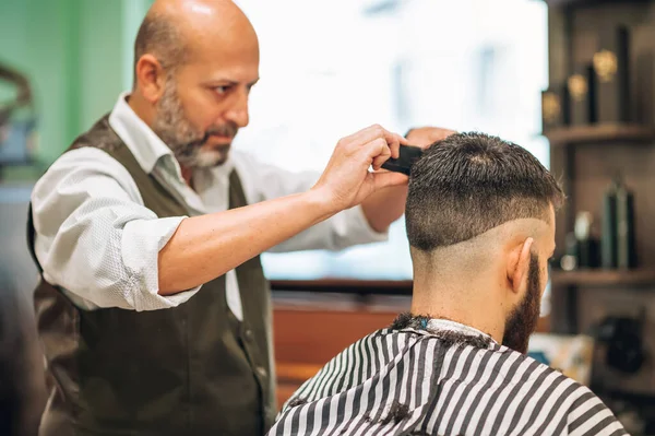 Aged bearded male barber with comb and scissors doing haircut to man in contemporary salon