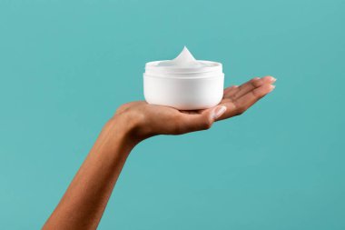 Tanımlanamayan Afro-Amerikalı kadın turkuaz arka planda beyaz plastik kavanoz ve vücut kremi gösteriyor.