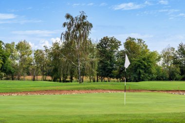 Golf sahasında büyüyen yeşil çimlerin, yemyeşil ağaçların ve bulutlu mavi gökyüzünün beyaz bayrağıyla görüntüsü.
