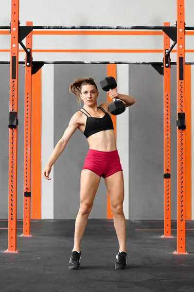 Tam boy, kaslı vücutlu, spor kıyafetleri içinde uzun saçlı, ağırlık kaldıran ve modern spor salonunda spor yaparken kameraya bakan kararlı genç bayan.