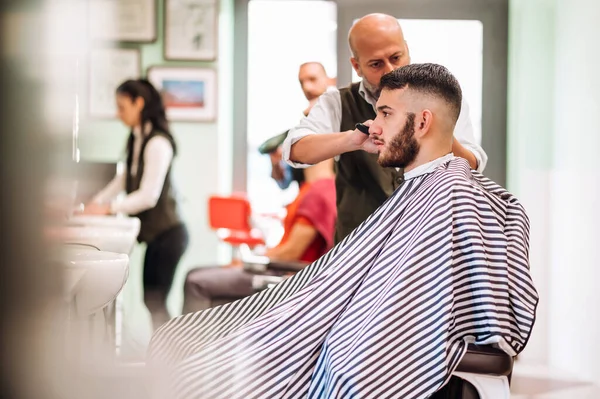 现代理发店穿着条纹斗篷 留着胡子的男性顾客坐在专业理发椅子上的侧视图 图库图片