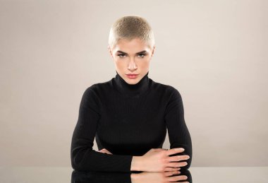 Siyah balıkçı yaka kazaklı, ciddi bir genç kadın portresi açık kahverengi arka planda kameraya bakarken masada çapraz el ele oturuyor ve yaslanıyor.